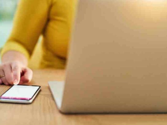 Person In A Yellow Sweater Using A Smartphone Next To An Open Laptop On A Wooden Table, Blurred Background.