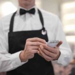 A Waiter In A White Shirt, Black Vest, And Bow Tie Holds A Notepad And Pen, Ready To Take An Order In A Restaurant Setting.