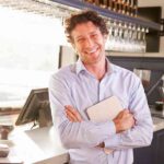 A Smiling Man In A Blue Shirt Standing With Crossed Arms In A Sunlit Bar, Holding A Tablet.