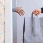 Two Hands Exchanging A Gray Shopping Bag Through A Doorway, Highlighting A Transaction Or Delivery.