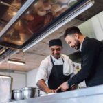 Two Men, One In Chef Attire And The Other In A Suit, Collaborating And Smiling In A Professional Kitchen.