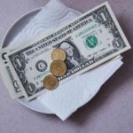 A One Dollar Bill And Several Coins On A White Paper Napkin Placed On A Plate, Possibly Symbolizing Economical Dining Or Budgeting Concept.